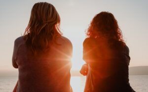 two woman sitting on beach sand while facing sunlight