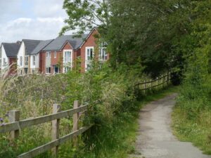 A dirt path leads past a wooden fence towards houses.